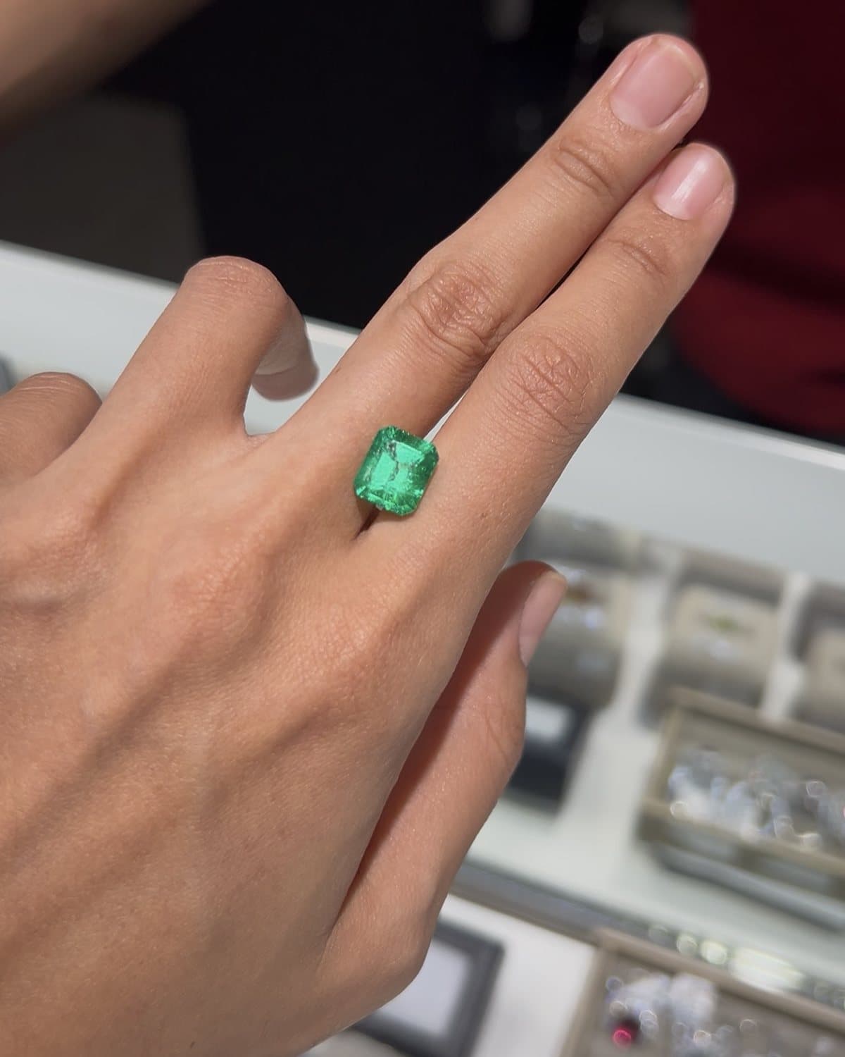 A large emerald-cut emerald resting between two fingers at a dealer's counter, trays of loose stones behind